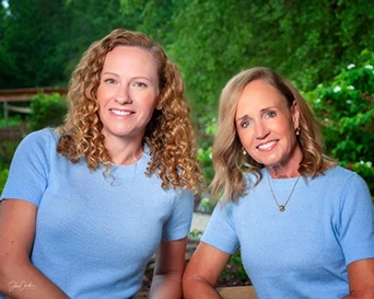 Two female Dentists wearing matching light blue tops smiling outdoors with lush green foliage backdrop